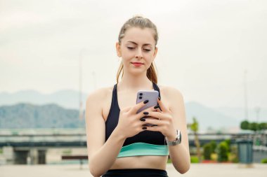 Şehir parkında spor kıyafetler giyen genç esmer kadın, açık havada telefon ekranına bakıyor ve telefon kullanıyor. Arkadaşlarla mesajlaşma, video izleme veya sosyal medyada kaydırma.