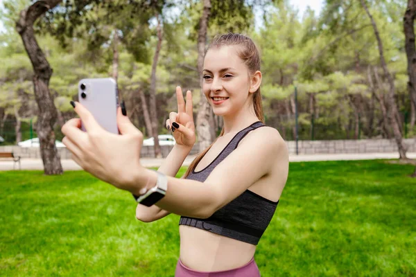 Cheerful brunette woman wearing sportive clothes on city park, outdoors smiling with happy face winking at the smart phone doing victory sign or gesture, showing peace. Number two.