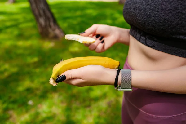Siyah spor sutyen giyen esmer kadın şehir parkında duruyor, elinde muz ve pirinç krakeri var. Açık hava sporu ve sağlıklı yaşam kavramları.