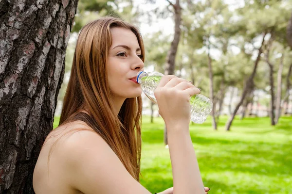 Kentin parkında duran spor sutyenini takan genç bir kadın. Yüzünde gülümsemeyle serinlemek için açık havada su içiyor. Açık hava sporları, sağlıklı yaşam kavramları..