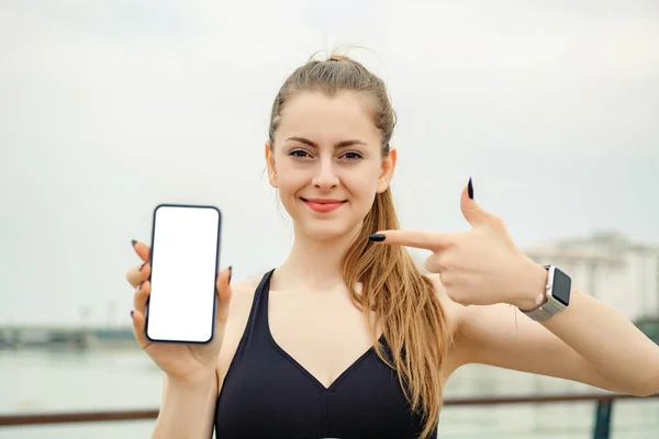 Şehir parkında spor kıyafetler giyen beyaz bir kadın, açık havada bir telefon tutuyor ve telefonda gülen yüzlü birini işaret ediyor. Cep telefonu ekranına bakıyor..