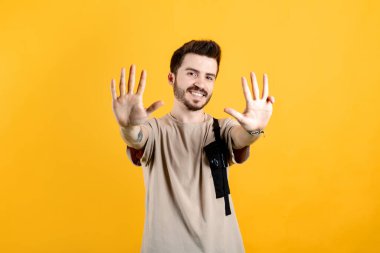 Sarı arka planda izole edilmiş bej tişört giyen genç adam gülümserken dokuz numarayı işaret ediyor..