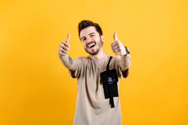 Sarı arka planda izole edilmiş bej tişört giyen bir adamın portresi büyük başarı için el hareketi yapıyor. İki elini de havaya sok..