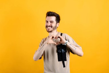 Sarı arka planda izole edilmiş bej tişört giyen, kendinden emin gülümseyen genç adam parmaklarıyla kalp figürünü gösteriyor. Sevgiyle gülümseyen, elleriyle kalp sembolü yapan. Romantik konsept.