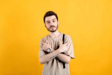 Sarı arka plan üzerinde izole edilmiş bej tişört giyen yakışıklı beyaz adam çapraz eller ve her iki taraftaki kopyalama alanını işaret ediyor.
