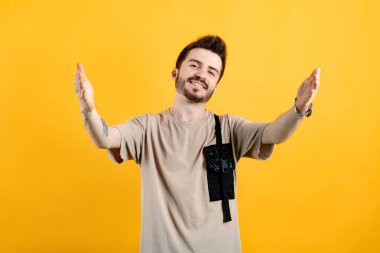 Sarı arka planda izole edilmiş bej tişört giyen kendine güvenen genç adam sarılmak için kollarını açmış gülümsüyor. Mutluluğu kucaklayan neşeli bir ifade..