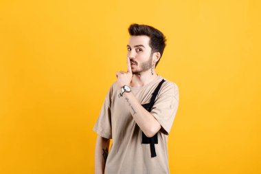 Sarı arka plan üzerinde izole edilmiş bej tişört giyen yakışıklı beyaz adam şşşş hareketi yapıyor, şaşırtıcı bir şekilde yana bakıyor, garip bir şey fark ediyor, koyu renk sakalı var..