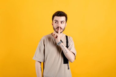 Beyaz tişörtlü beyaz bir adam sarı arka planda izole bir şekilde poz veriyor. Şşş hareketi yapıyor. Şaşırtıcı bir şekilde, garip bir şey fark ediyor..