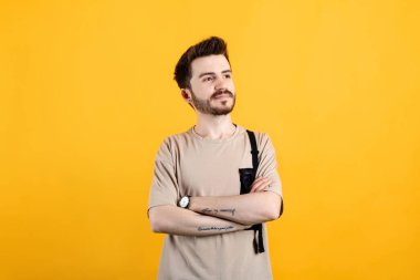 Sarı arka planda izole edilmiş bej tişört giyen yakışıklı beyaz adam kollarını kavuşturuyor ve gülümsüyor..