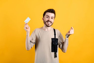 Tişört giyen neşeli genç adam sarı arka planda izole bir şekilde poz veriyor. Elinde kredi kartı ve para işareti, gülen surat ve mutlu kazançlar var.
