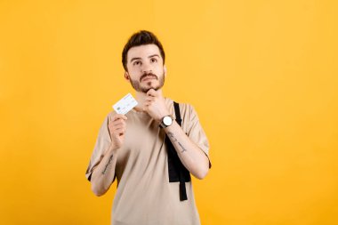 Sarı arka planda izole edilmiş bej tişört giyen, kendine güvenen genç bir adam kredi kartı ya da banka kartı taşıyor soru soran ciddi bir yüz, çok karışık bir fikir..