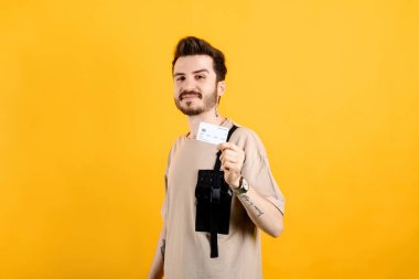 Sarı arka planda izole edilmiş bej tişört giyen bir adamın portresi. Kameraya bakarken plastik kredi kartı ya da banka kartı taşıyor..
