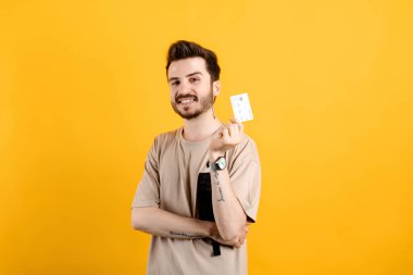 Sarı arka planda izole edilmiş bej tişört giyen mutlu genç adam kameraya bakarken plastik kredi kartı ya da banka kartı tutuyor..