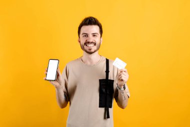 Sarı arka plan boş ekran ve kredi kartı üzerinden izole edilmiş bej tişört giyen, kendine güvenen genç adam, alışveriş veya çevrimiçi ödeme, bankacılık, modelleme için en yeni mobil uygulamayı tavsiye ediyor.