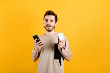 Beyaz tişört giyen beyaz bir adam sarı arka planda izole edilmiş bir şekilde poz veriyor. Elinde kredi kartı ve hayal kırıklığına uğramış bir akıllı telefon, omuz silkme, iflas konsepti var.