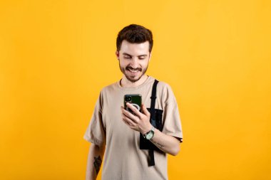 Sarı arka planda izole edilmiş bej tişört giymiş, kendine güvenen genç adam akıllı telefon ekranlarına bakıyor, web sayfalarına bakıyor ve arkadaşlarıyla sohbet ederken gülümsüyor..