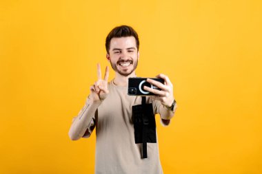Sarı arka planda izole edilmiş bej tişört giyen mutlu genç adam zafer işareti yapıyor ve selfie çekerken gülümsüyor. Video çağrısı veya sosyal medya kavramları.