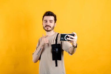 Sarı arka planda izole edilmiş bej tişört giyen yakışıklı beyaz adam cep telefonunda selfie çekerken işaret parmağıyla işaret ediyor. İletişim kavramları.