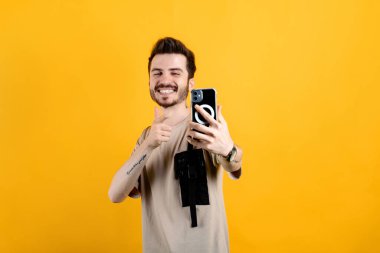 Sarı arka planda izole edilmiş bej tişört giyen mutlu genç adam cep telefonuna işaret parmağı yerleştirirken büyük gülümsemeyle selfie çekiyor. İletişim kavramları.