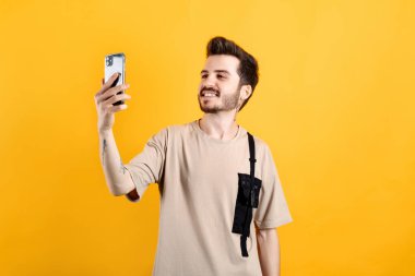 Sarı arka planda izole edilmiş bej tişört giyen yakışıklı beyaz adam büyük gülümsemeli telefon ekranına bakarak selfie çekiyor..