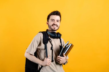 Bej tişört giyen bir adamın portresi sarı arka plan öğrencisinin üzerinde izole edilmiş kitaplar ve sırt çantasıyla kameraya bakıyor. Lise üniversite konsepti.