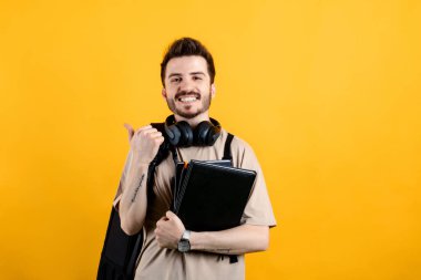 Sarı arka planda izole edilmiş bej tişört giyen yakışıklı beyaz adam gülümsüyor ve başparmağıyla yan tarafı işaret ediyor..