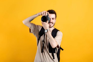 Beyaz tişört giyen beyaz bir adam sarı arka planda izole edilmiş bir şekilde DSR kamerayla fotoğraf çekiyor. Fotoğrafçı yüzünü kamerayla kapatıyor..