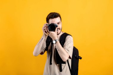Sarı arka planda izole edilmiş rahat kıyafetler giyen yakışıklı genç adam DSR kamerasıyla fotoğraf çekiyor. Fotoğrafçı yüzünü kamerayla kapatıyor..
