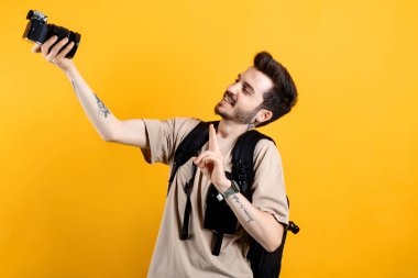 Sarı arka planda izole edilmiş bej tişört giyen genç adam profesyonel bir DSLR kamerasıyla selfie çekiyor..