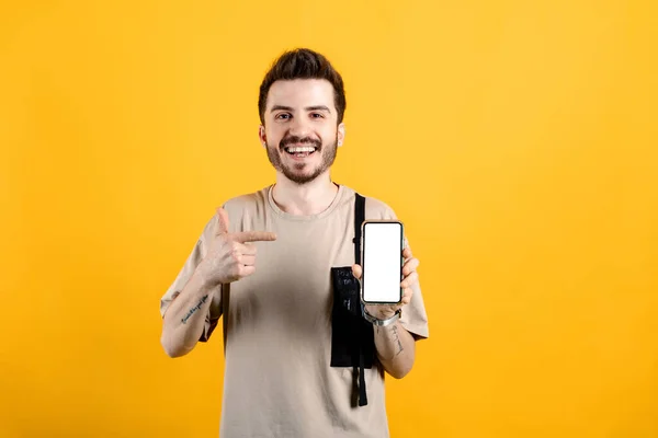 Happy casual man wearing t-shirt posing isolated over yellow background showing to camera and pointing at device. Gadget with empty free space for mock up. White blank screen.