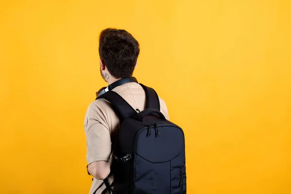Handsome caucasian man wearing beige t-shirt posing isolated over yellow background student wearing a backpack with headphones around his neck. Back view photo.