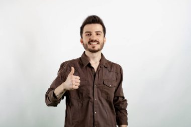 Beyaz arka planda izole edilmiş rahat kıyafetler giyip mutlu bir şekilde el hareketleriyle gülümseyen genç bir adam. Kameraya bakıp başarıyı gösteren ifade onaylanıyor.