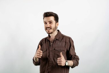 Beyaz arka planda izole edilmiş tişört giyen mutlu genç adam kameraya gülümsüyor ve başparmağını kaldırıyor. Kameraya bakıp başarıyı gösteren ifade onaylanıyor.