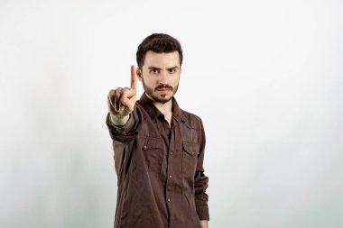 Kahverengi gömlekli genç adam beyaz arka planda yalnız poz veriyor. Parmağını kaldırmış ve kızgın bir ifadeyle işaret ediyor..
