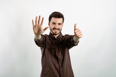 Kahverengi gömlekli neşeli genç adam beyaz arka planda izole edilmiş bir şekilde poz veriyor ve gülümserken altı numarayı işaret ediyor..