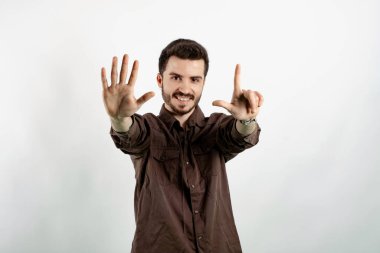 Kahverengi gömlekli neşeli genç adam beyaz arka planda izole edilmiş bir şekilde poz veriyor ve gülümseyerek yedi numaralı parmaklarıyla işaret ediyor..