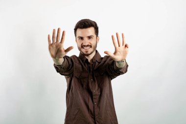 Kahverengi gömlekli neşeli genç adam beyaz arka planda izole edilmiş bir şekilde poz veriyor ve dokuz numaralı parmaklarıyla gülümseyerek işaret ediyor..