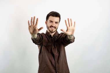 Kahverengi gömlekli neşeli genç adam beyaz arka planda izole edilmiş bir şekilde poz veriyor ve gülümseyerek on numaralı parmaklarıyla işaret ediyor..