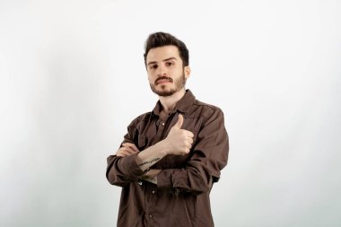Kahverengi gömlekli genç adam beyaz arka plan kolları üzerinde izole bir şekilde duruyor, kameraya bakıyor, gülümsüyor, başparmak hareketi yapıyor.