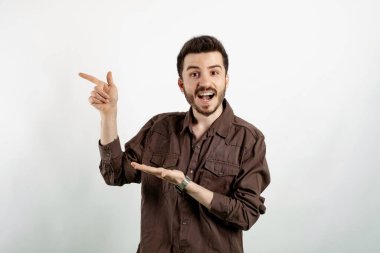 Kahverengi gömlekli neşeli genç adam beyaz arka planda yalnız poz veriyor. Gülümsüyor ve boş avucunu işaret ediyor..