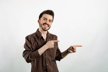 Kahverengi gömlekli neşeli beyaz adam beyaz arka planda yalnız poz veriyor gülümsüyor ve kameraya bakıyor iki eliyle ve parmaklarıyla yandan işaret ediyor..