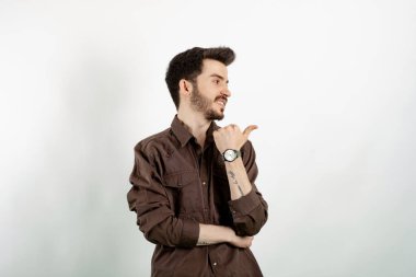 Yakışıklı, günlük kıyafetler giyen, beyaz arka planda izole edilmiş bir şekilde poz veren ve baş parmağıyla yan tarafı gösteren genç bir adam..