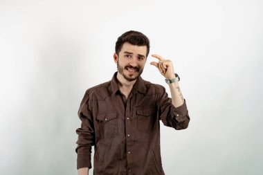 Beyaz arka planda izole edilmiş günlük giysiler giymiş, kendinden emin gülümseyen genç bir adam parmaklarıyla ölçü alırken kameraya bakıyor. Acıyarak küçük boy uzunluğu gösteriyor.