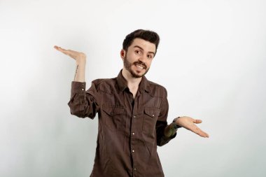 Yakışıklı, sıradan kıyafetler giyen, beyaz arka planda yalnız poz veren genç adam omuzlarını silkeliyor, şaşkın görünüyor, ellerini yana açıyor, bilemiyorum, belli olmuyor, duruyor.