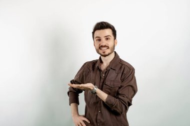 Kahverengi gömlekli genç adam beyaz arka planda yalnız poz veriyor. Neşeli bir şekilde gülümsüyor, mutlu hissediyor ve fotokopi alanında avuç içiyle bir konsept gösteriyor.