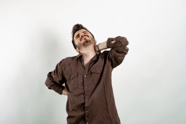 Kahverengi gömlekli genç adam, beyaz arka planda izole bir şekilde poz veriyor. Çabaladığı için sırt ağrısı çekiyor. Boynuna ve beline dokun..