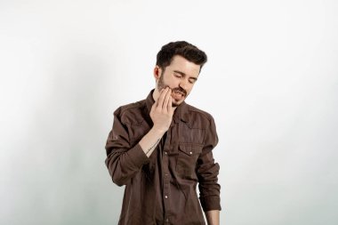 Beyaz arka planda izole edilmiş rahat kıyafetler giyen yakışıklı genç adam acı hissediyor, yanağını eliyle tutuyor, kötü diş ağrısından muzdarip. Acı verici bir ifade..