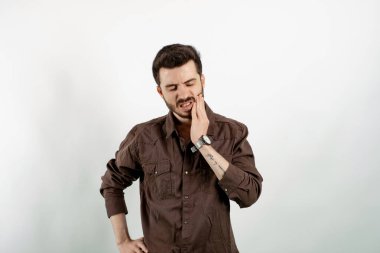 Kahverengi gömlekli genç bir adamın portresi beyaz arka planda izole edilmiş bir şekilde duruyor acıyı hissediyor, yanağını eliyle tutuyor, kötü diş ağrısından muzdarip. Acı verici bir ifade..