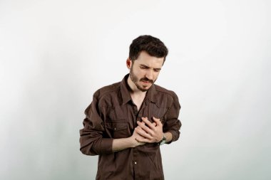 Beyaz arka planda yalnız poz veren beyaz giysili yakışıklı beyaz adam ellerini göğsüne bastırıyor ve kalp krizi geçiriyor..