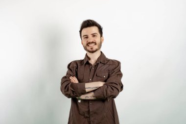 Kahverengi gömlekli neşeli beyaz bir adam beyaz arka planda yalnız poz veriyor. Kameraya bakarak kollarını kavuşturuyor. Pozitif kişi.